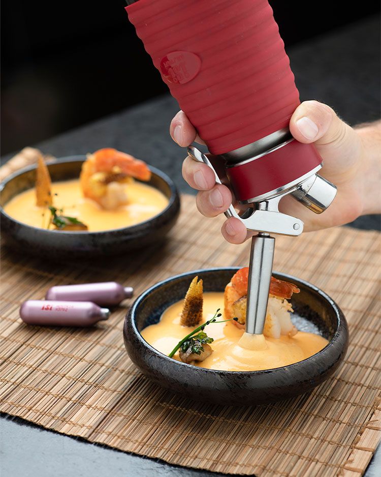 Jemand sprüht mit einem iSi Gourmet Whip Tom Kha Gai Suppe in eine Schüssel. im Hintergrund eine bereits gefüllte Schüssel und iSi Charge Kapseln.