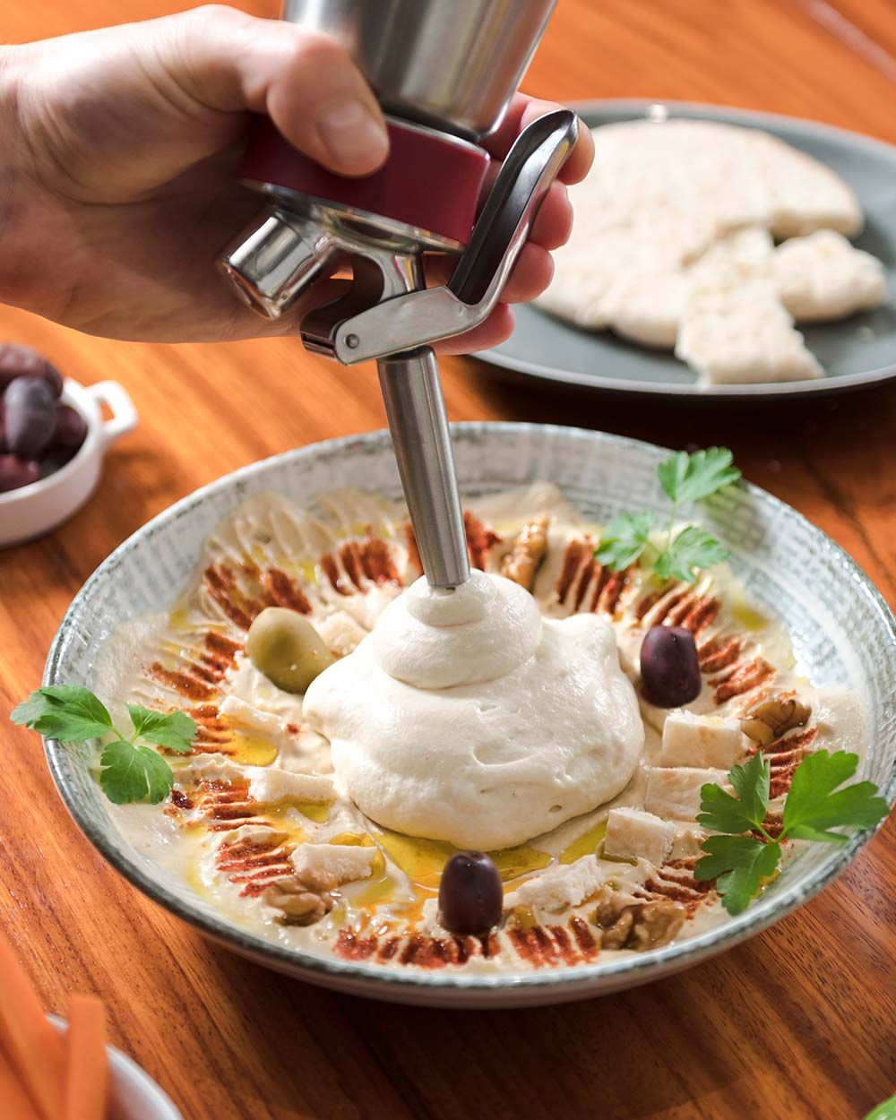 Jemand sprüht veganen Hummus in eine Schüssel, im Hintergrund Pita-Brot. 