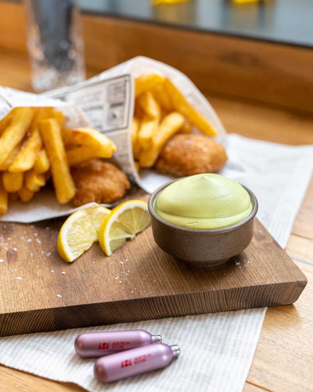 Zwei Tüten mit Fish and Chips, davor  eine Schüssel mit Erbsen Espuma und zwei iSi Charge Kapseln. 