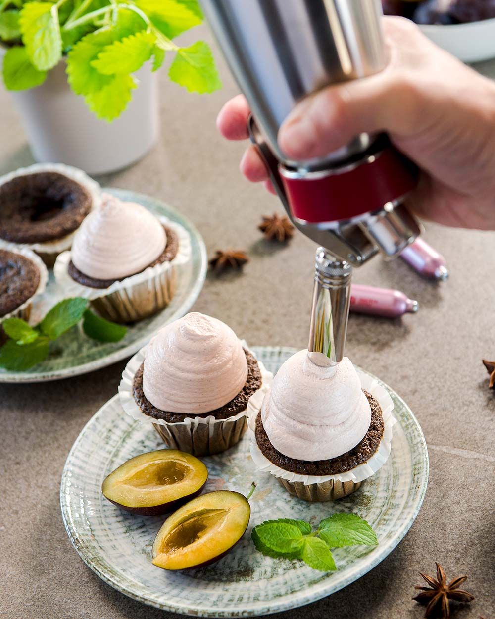 Jemand sprüht mit einem iSi Gourmet Whip Cupcake Topping auf Mohn-Pflaumen Cupcakes, daneben weitere Pflaumen und iSi Charge Kapseln. 