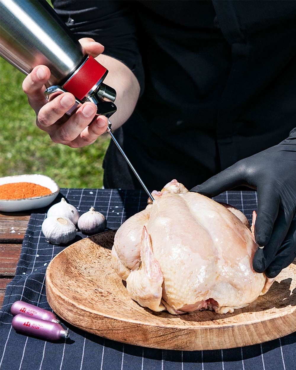 Jemand benutzt ein iSi Gourmet Whip um ein ganzes Huhn zu marinieren. Daneben Gewürze, Knoblauch und zwei iSi Charge Kapseln.
