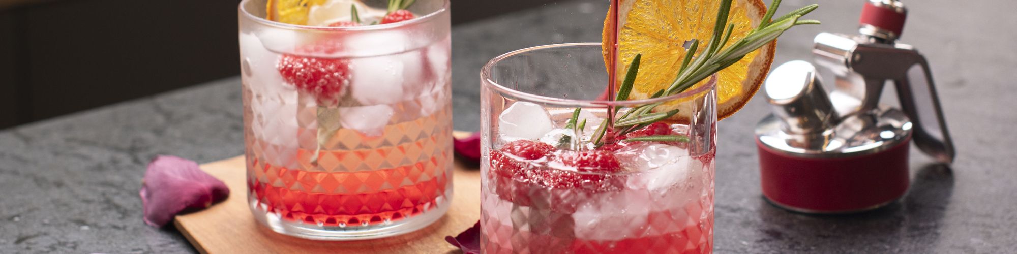 Zwei Cocktailgläser mit roten Drinks, Eiswürfeln, Himbeeren, Orangenscheiben und Rosmarin stehen auf einem Tisch, daneben liegt ein iSi Gerätekopf.