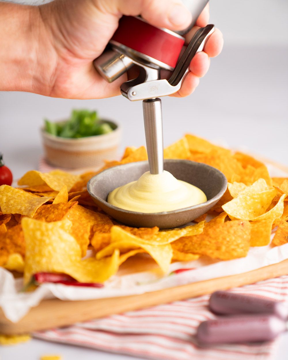 Jemand sprüht mit einem iSi Gourmet Whip Käse-Espuma in eine Schüssel, rundherum liegen Nachos. Im Vordergrund zwei iSi Charge Kapseln. 