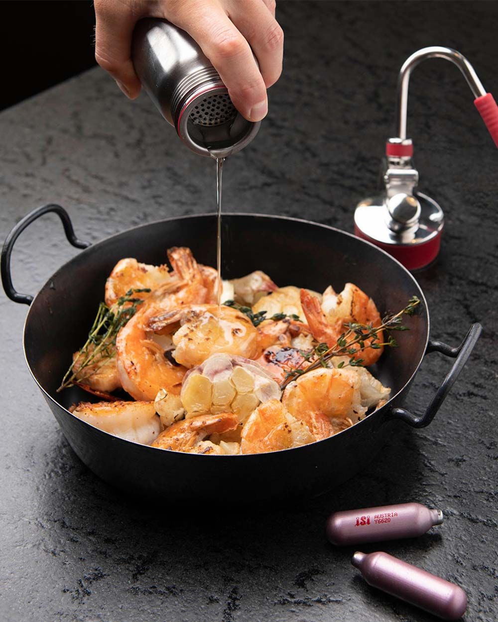 Jemand gießt Gartenkräuteröl aus einem iSi Gourmet Whip in eine Pfanne mit Meeresfrüchten.  Davor liegen zwei iSi Charge Kapseln. 