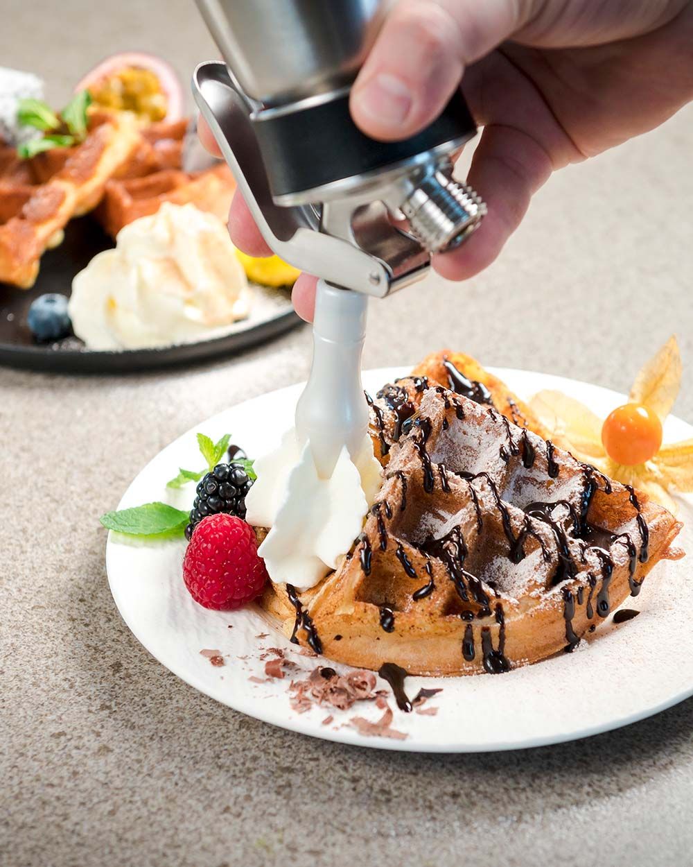 Auf dem Bild wird mit einem iSi Sahnespender frische Schlagsahne neben eine mit Schokolade garnierte Waffel mit Beeren auf einem Teller gespritzt.