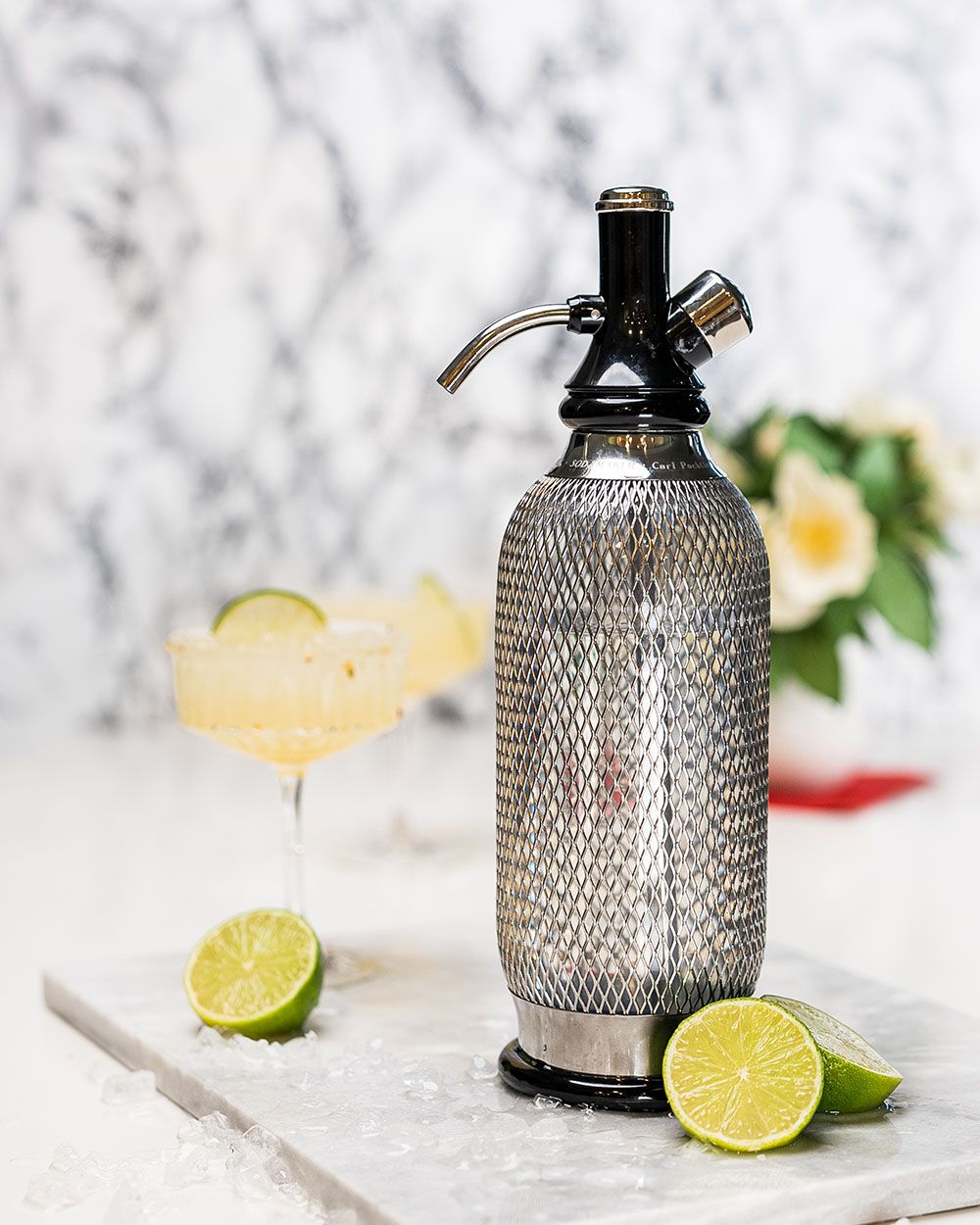 Ein iSi Sodamaker, daneben Limetten und ein Glas mit Holunder-Minz-Limonade. 