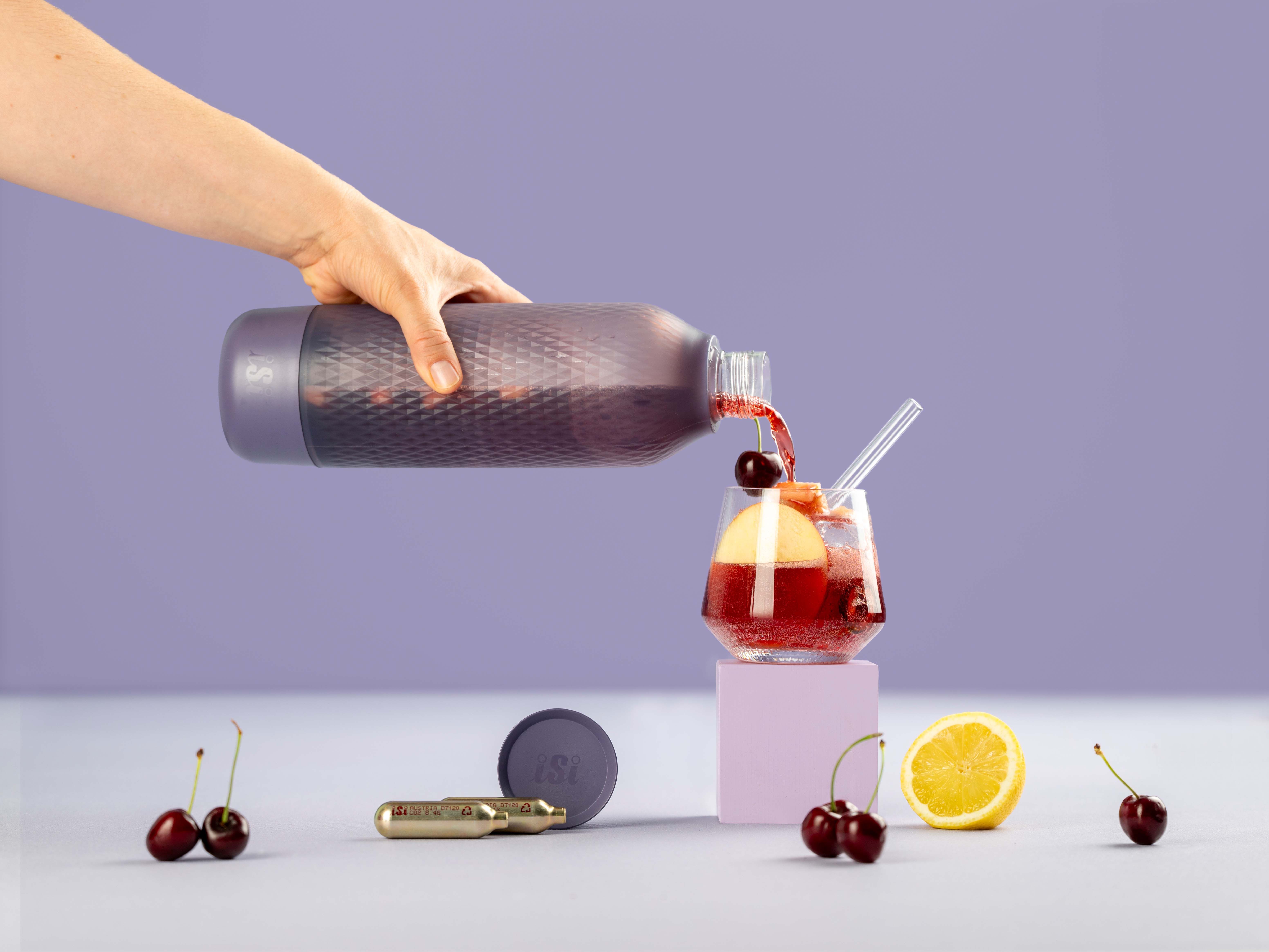 Jemand gießt Cherry Apple Tea aus einem iSi Twist'n Sparkle in ein Glas mit Eis und Früchten, daneben iSi Charge Kapseln und Früchte. 