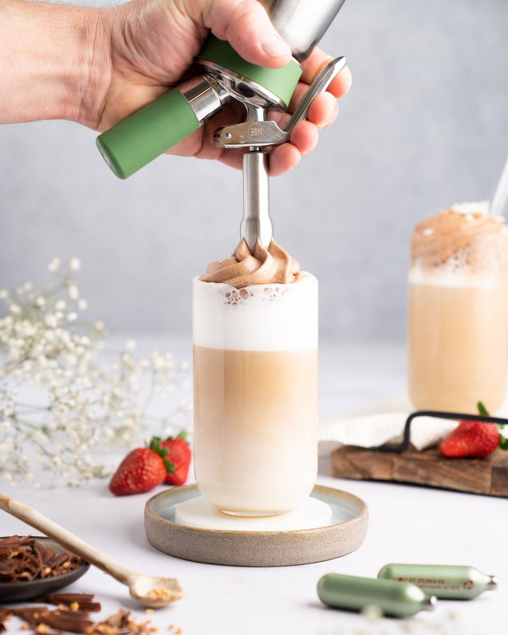 Jemand sprüht mit einem iSi Creative Whip Nutella-Topping auf ein Kaffeegetränk. Daneben zwei iSi Charge Kapseln, Erdbeeren und ein fertiges Getränk.