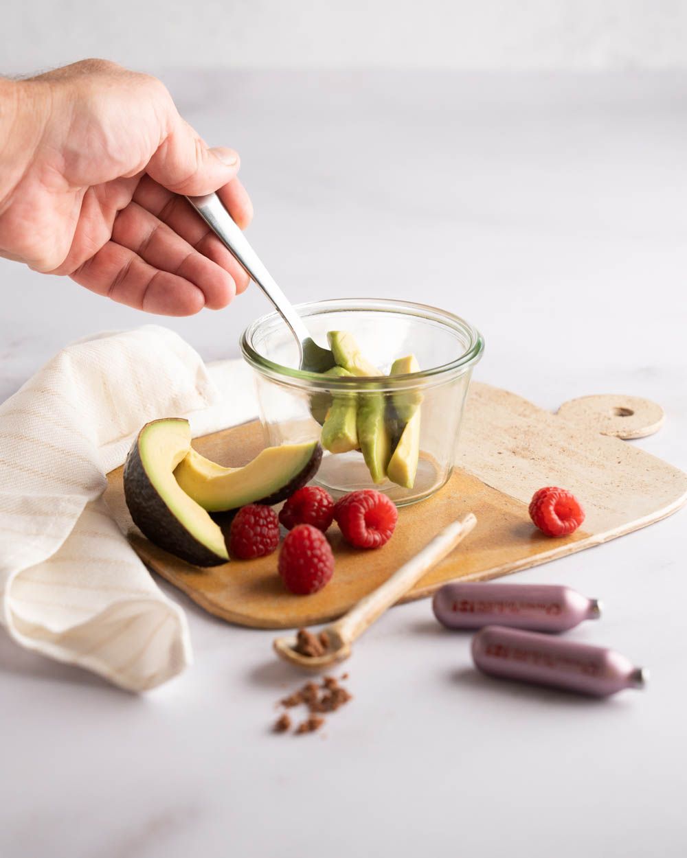 Auf dem Bild sieht man ein Glas mit Avocadostücken, daneben eine halbe Avocado, Himbeeren, zwei iSi Kapseln, ein Tuch und eine Hand mit einem Löffel.