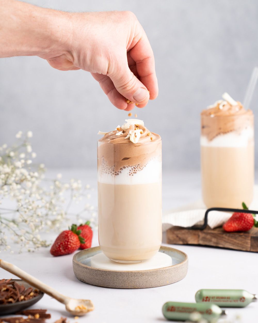 Jemand dekoriert ein Kaffeegetränk mit Nutella-Topping. Daneben Erdbeere, iSi Charge Kapseln und ein fertiges Getränk. 