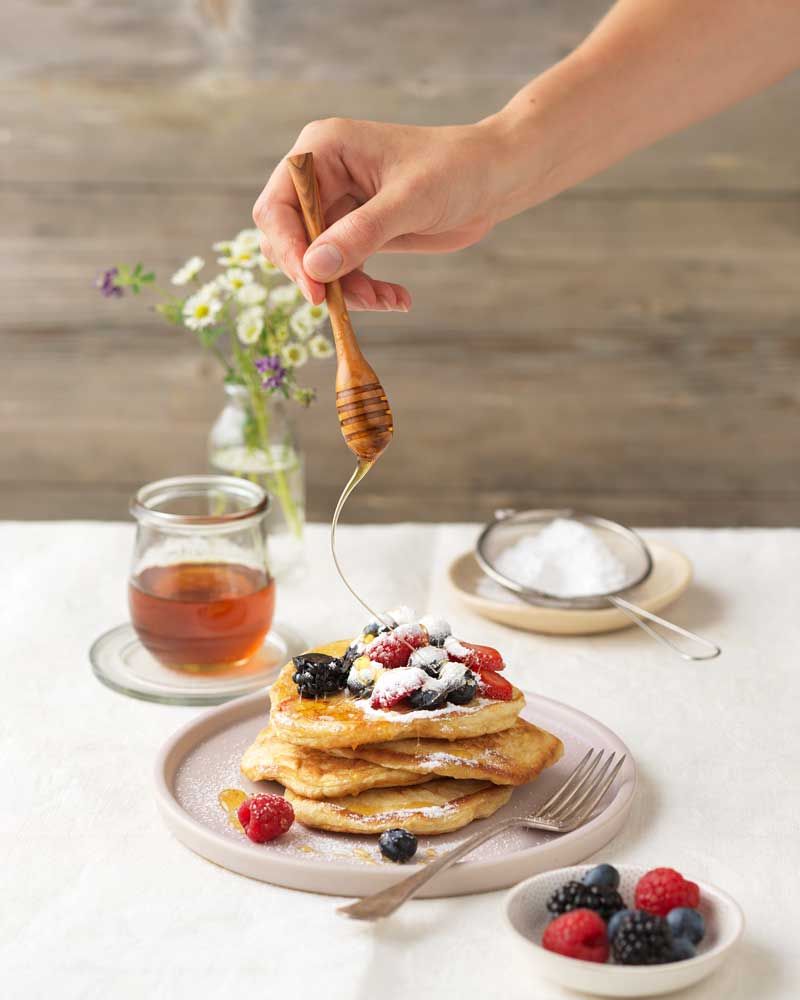 Jemand gibt Honig auf Pancakes mit Beere, davor mehr Beeren, Tee und Puderzucker. 