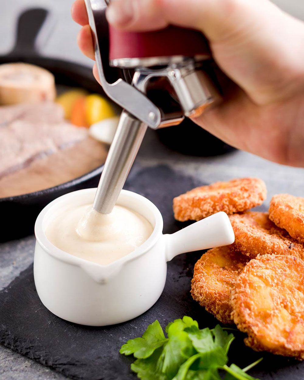 Jemand nutzt einen iSi Gourmet Whip um Schnittlauchsauce in eine Schüssel zu sprühen, daneben liegen Kartoffel-Laibchen. 