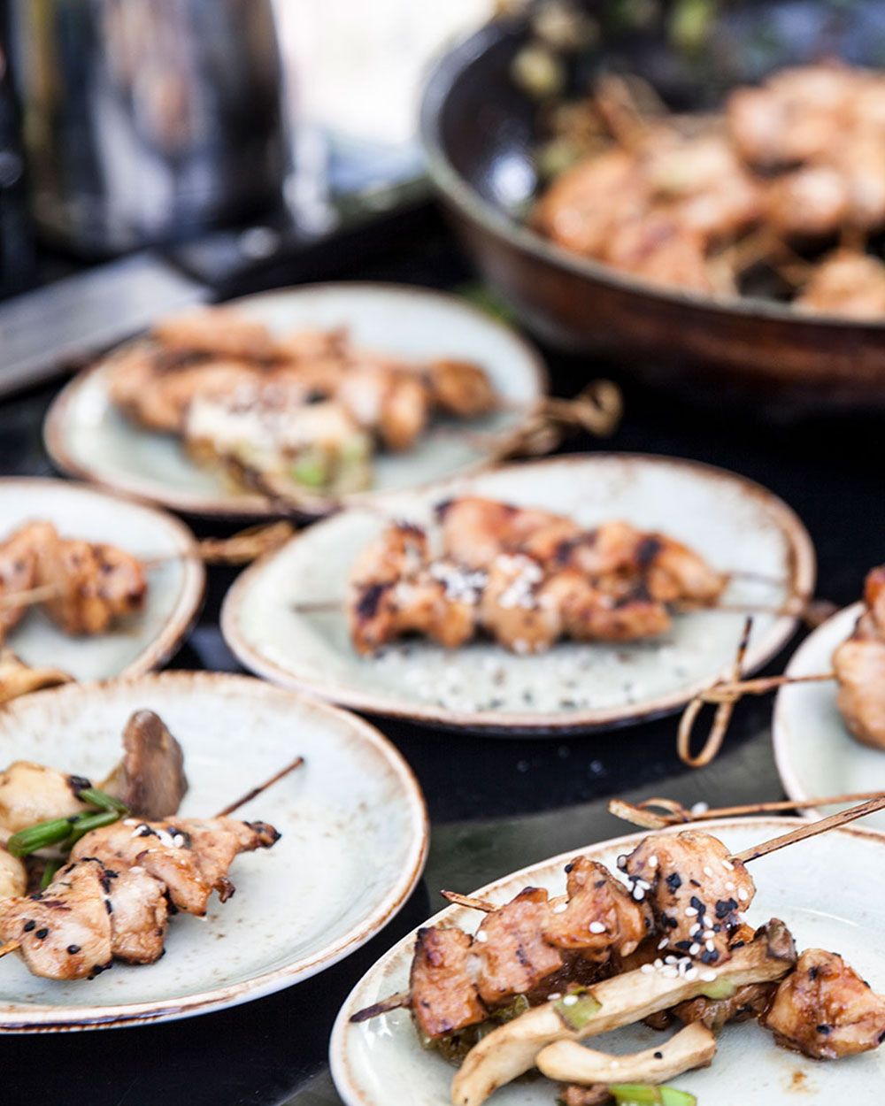 Auf dem Bild sind mehrere Teller mit gegrillten Hähnchenspießen zu sehen, garniert mit Sesam und serviert mit Pilzen. Im Hintergrund steht eine Pfanne mit weiteren Spießen.
