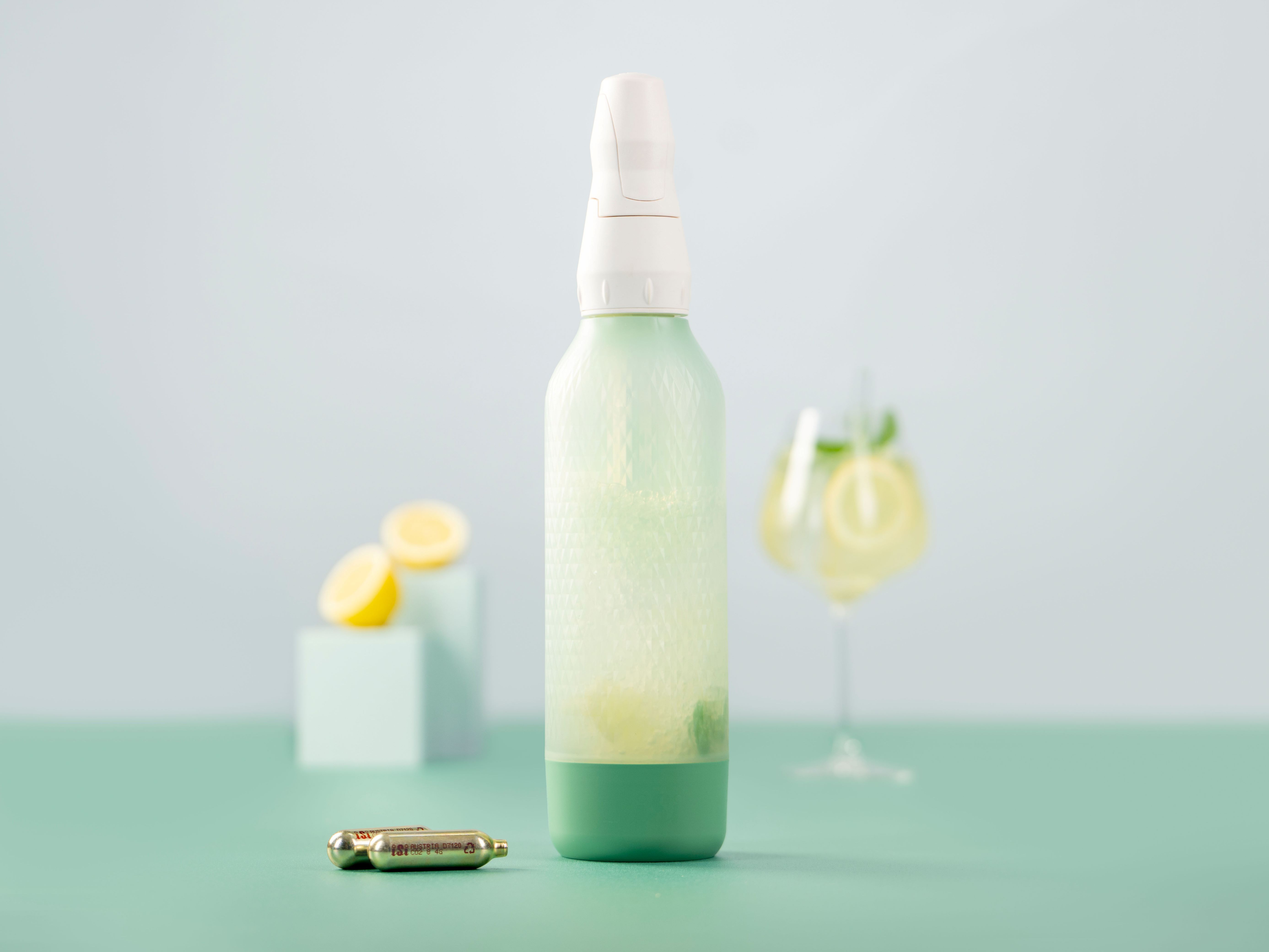 Ein iSi Twist'n Sparkle mit dem Getränk Hugo Bliss, daneben iSi Charge Kapseln, im Hintergrund Zitronen und ein Glas mit Hugo Bliss. 