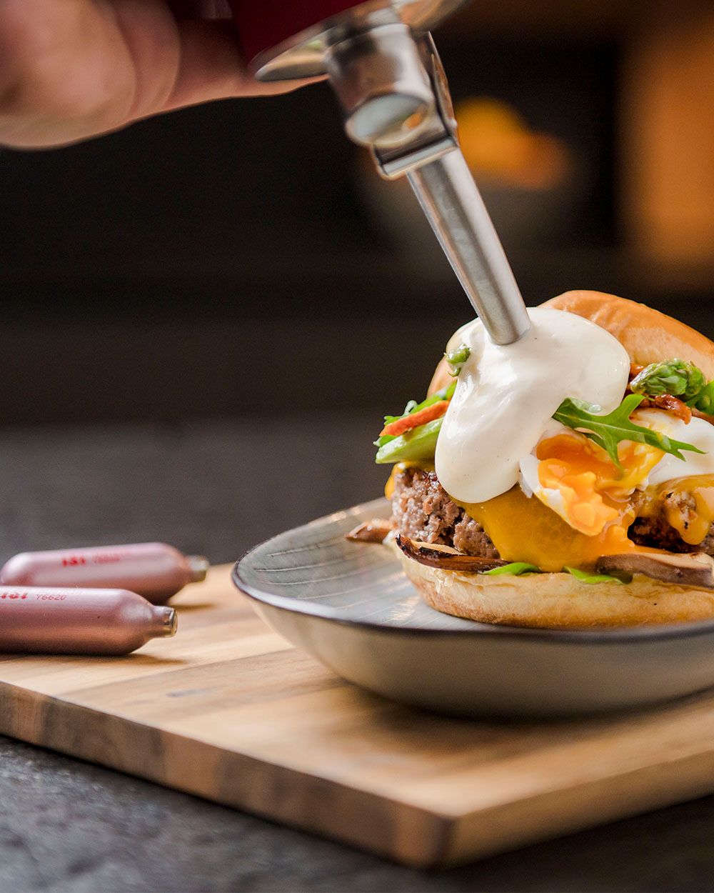 Jemand sprüht mit einem iSi  Whip Trüffel-Sauce-Hollandaise auf einen Burger, daneben iSi Charge Kapseln.