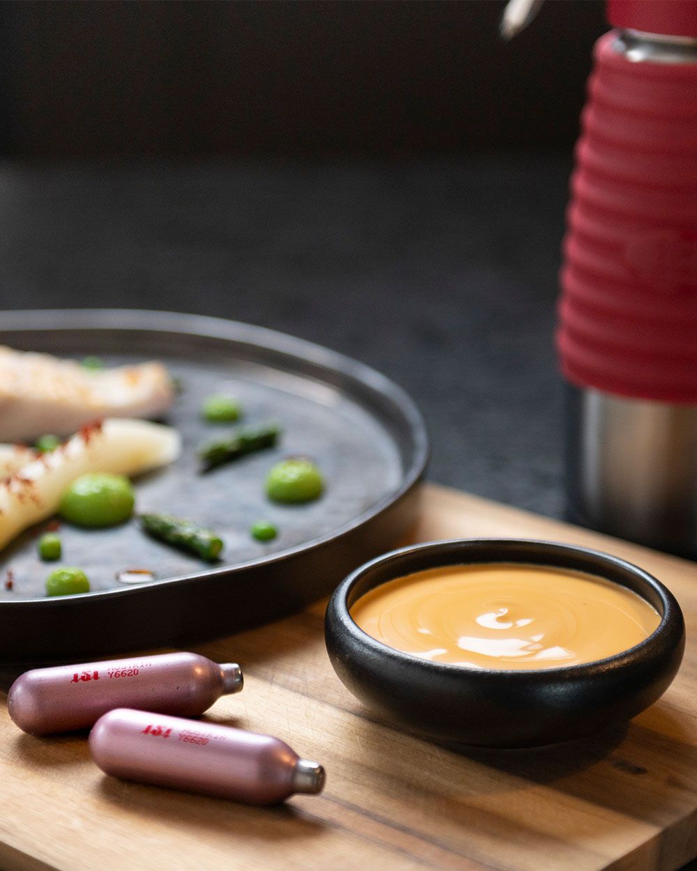 Auf dem Bild sieht man eine schwarze Schale mit heller Sauce, daneben ein Teller mit Gemüse und einen iSi Gourmet Whip.