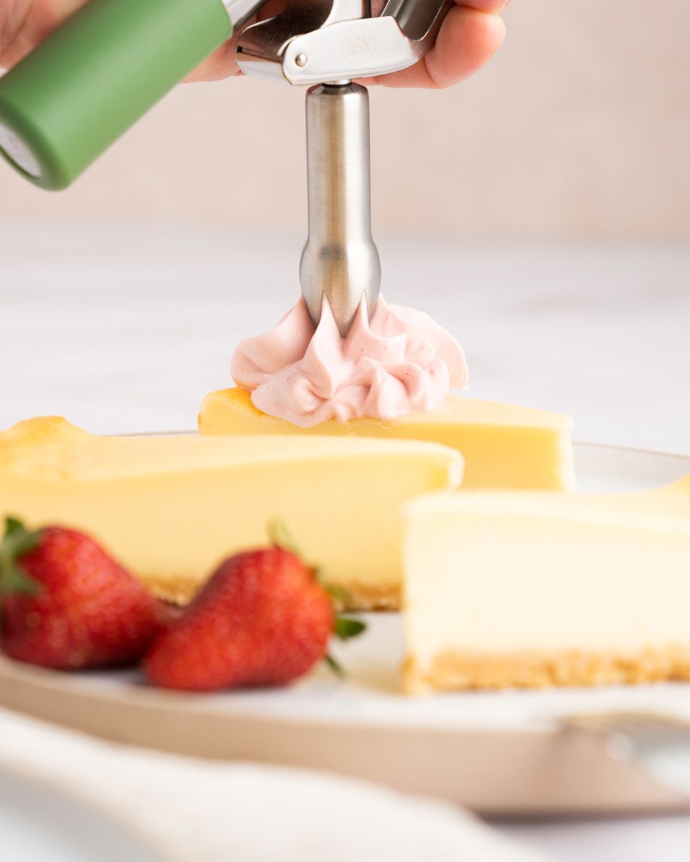 Jemand sprüht Erdbeer-Topping auf einen Cheesecake, daneben mehr Cheesecake und Erdbeeren. 