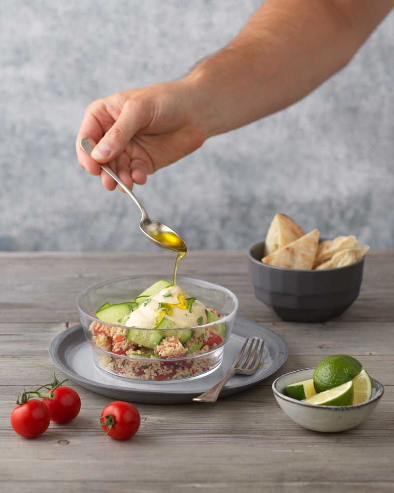 Jemand gießt Öl auf einen Salat, daneben sind Tomaten, Limetten und Brot. 