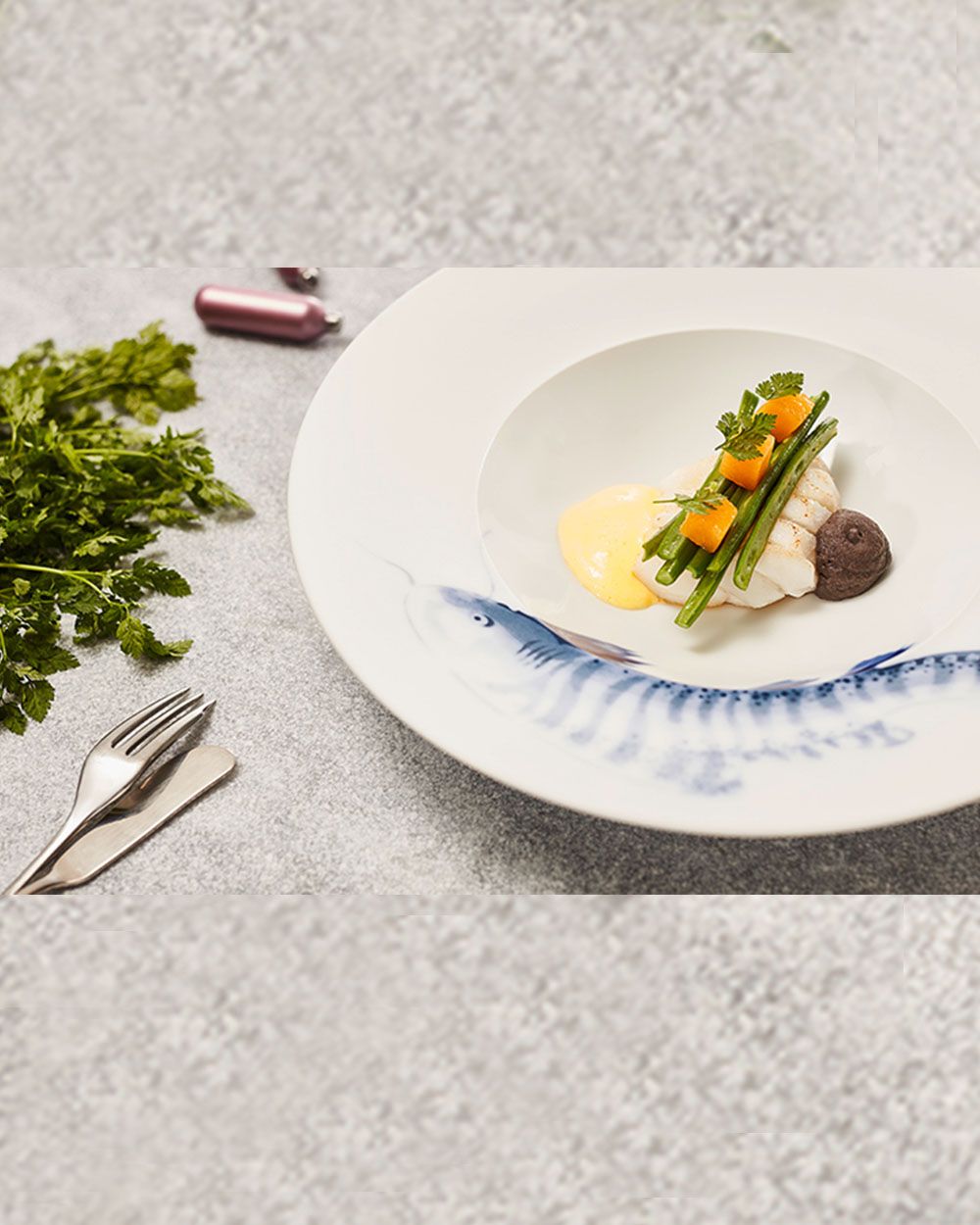 Auf dem Bild sieht man ein Fischfilet mit Gemüse, gelber Sauce und einem Klecks brauner Creme auf einem Teller mit Fischmotiv.