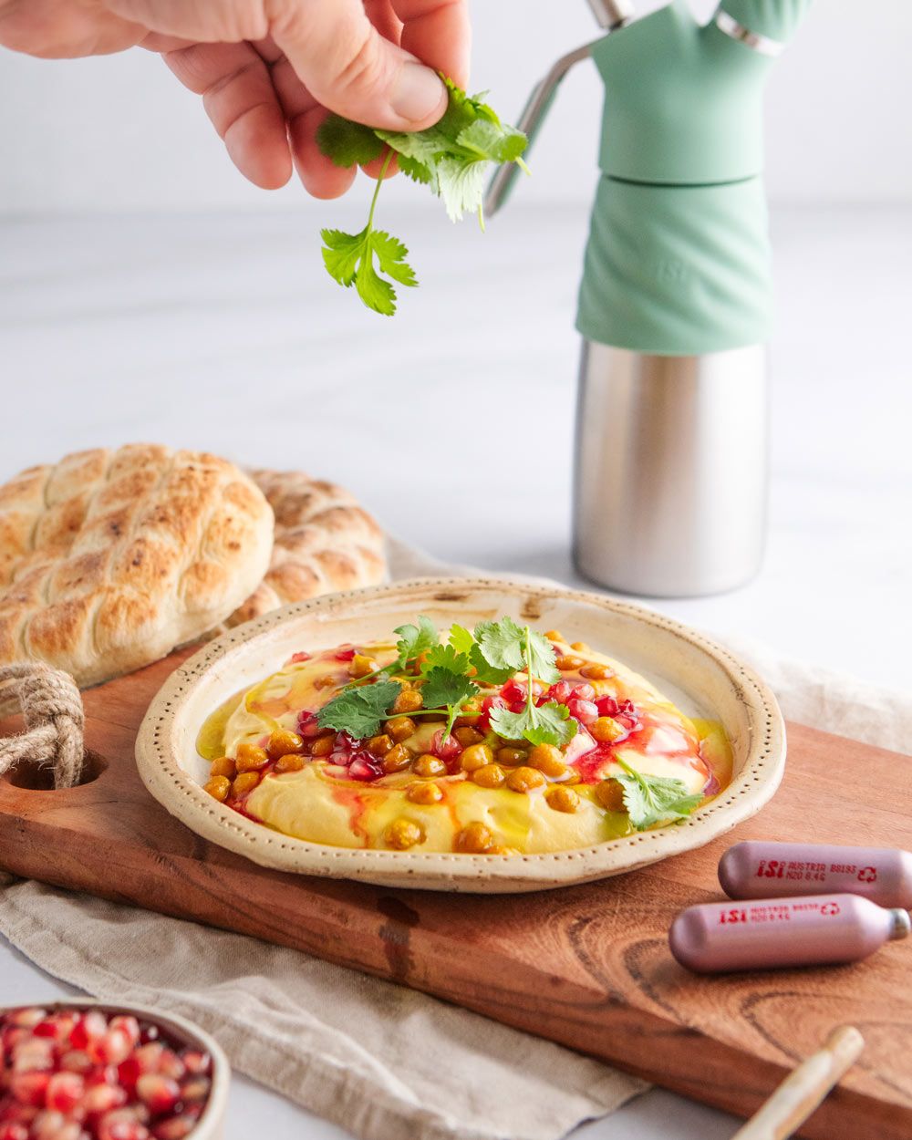 Jemand gibt Petersilie auf einen Teller mit Kürbishummus, daneben Pitabrot und zwei iSi Charge Kapseln. Im Hintergrund ein iSi Creative Whip.