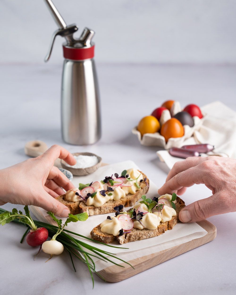 Zwei Personen greifen nach Broten mit Eiaufstrich und Kräutern. Daneben Radieschen, Schüsseln mit Gewürzen und im Hintergrund ein iSi Gourmet Whip, bunte Eier und zwei iSi Charge Kapseln. 