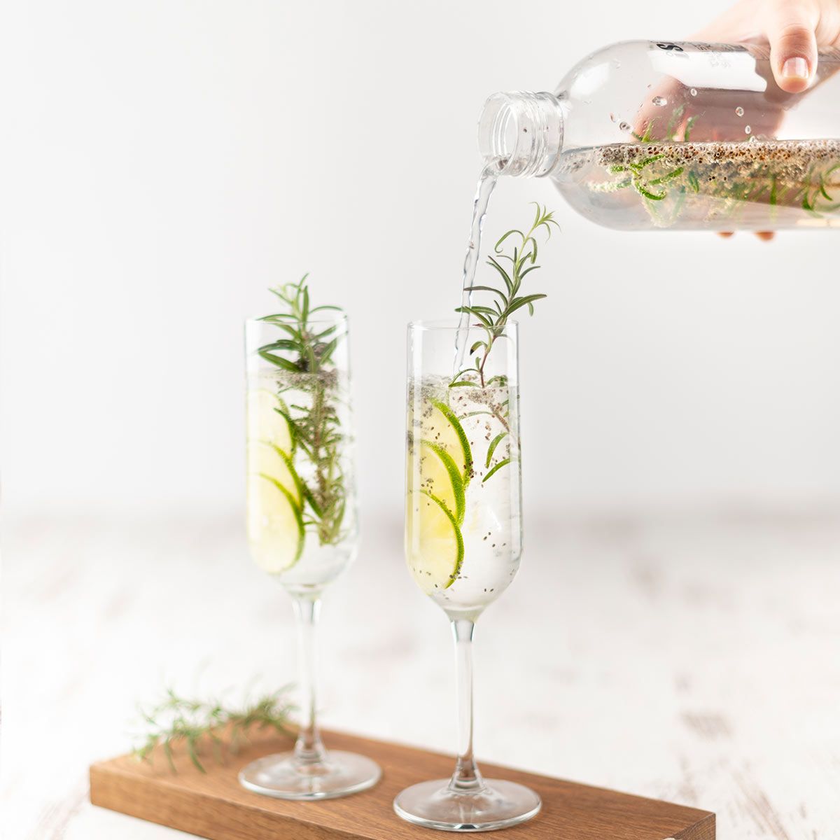 Eine Hand gießt sprudelndes Wasser aus einer Flasche mit Kräutern in zwei Gläser, die mit Limettenscheiben, Rosmarin und Eiswürfeln gefüllt sind.