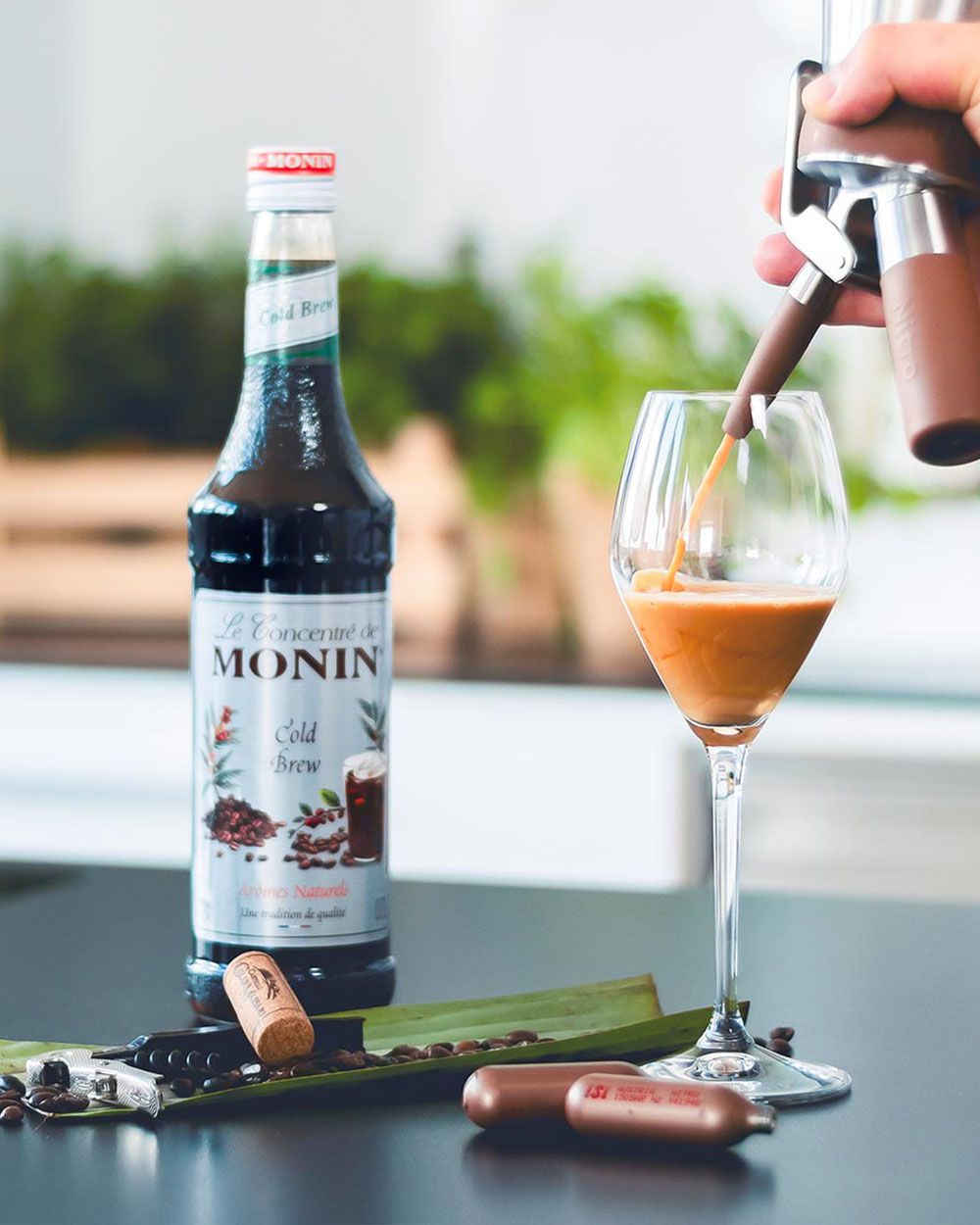 Jemand sprüht mit einem iSi Nitro ein Getränk in ein Glas, daneben iSi Charge Kapseln, Kaffeebohnen und Monin Sirup.