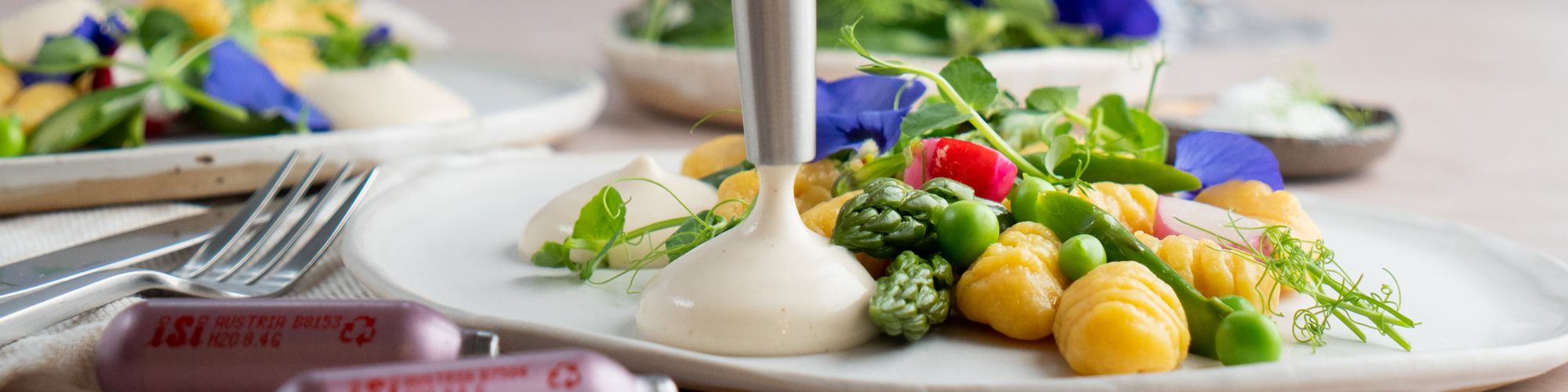 Ein Teller mit Gnocchi, grünem Gemüse und essbaren Blüten wird mit einer hellen Sauce aus einem iSi Whip verfeinert.