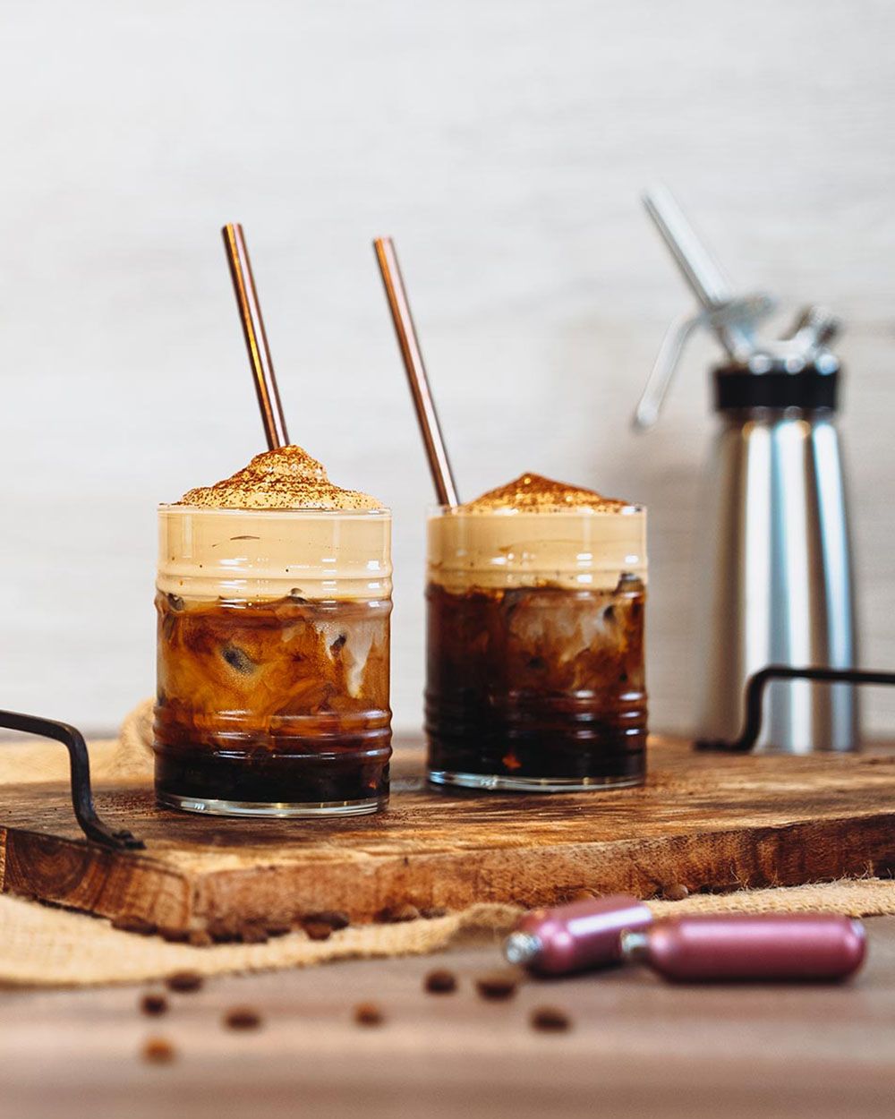 Zwei Gläser mit Eiskaffee und cremigem Schaumtopping stehen auf einem Holzbrett mit Metallstrohhalmen, im Hintergrund ist ein iSi Whip zu sehen und im Vordergrund liegen iSi Kapseln sowie Kaffeebohnen.