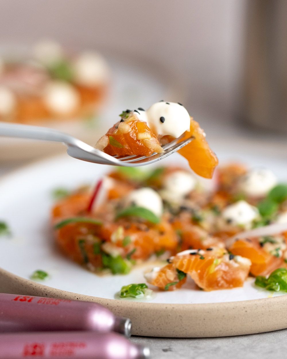 Jemand sticht mit einer Gabel einen Bissen Lachs Tatar mit Yuzu Creme von einem Teller. 