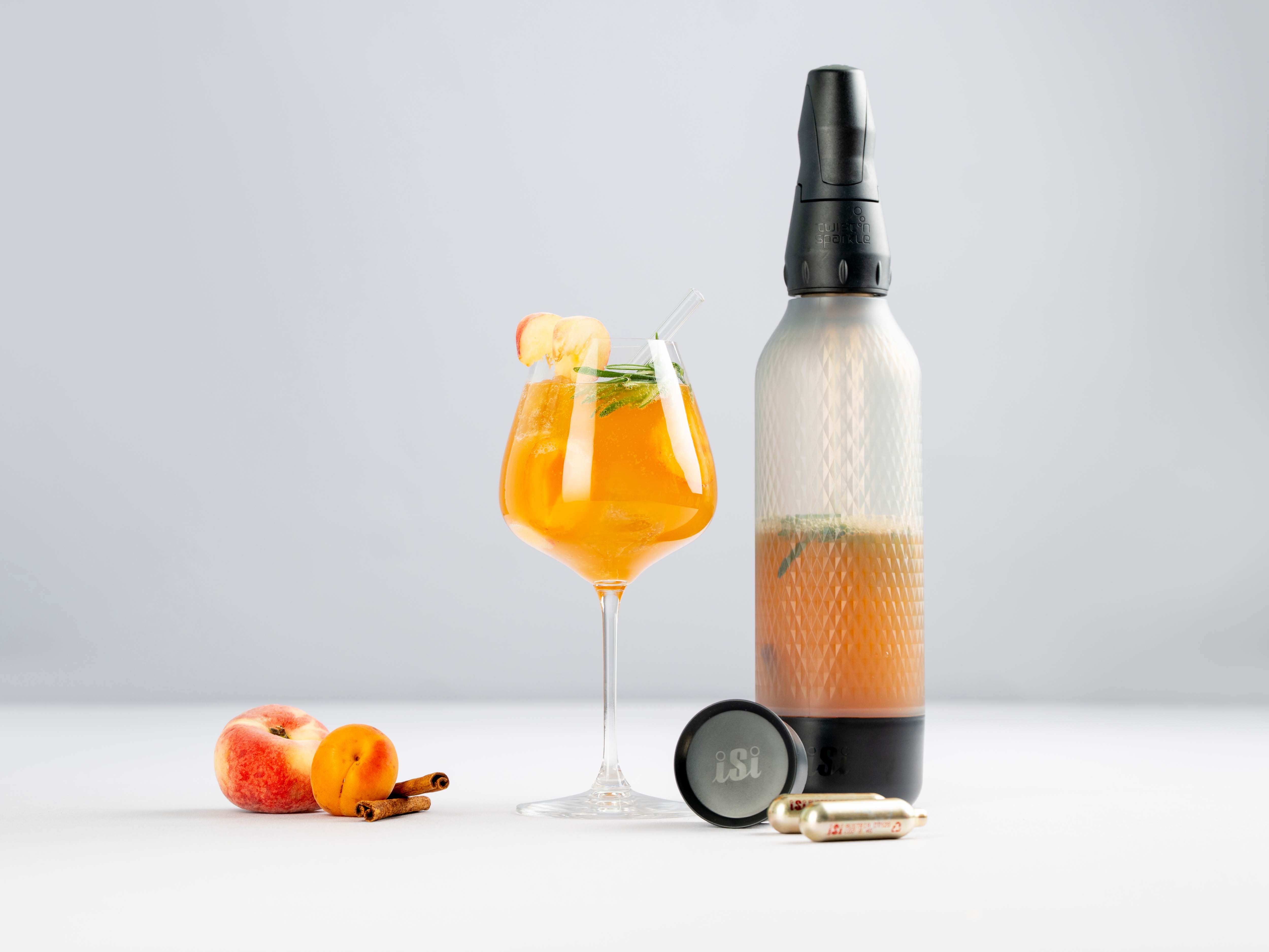 Ein Glas mit einem orangenen Getränk – Apricot Spritz – daneben Mandarinen und ein iSi Twist'n Sparkle und iSi Charge Kapseln.