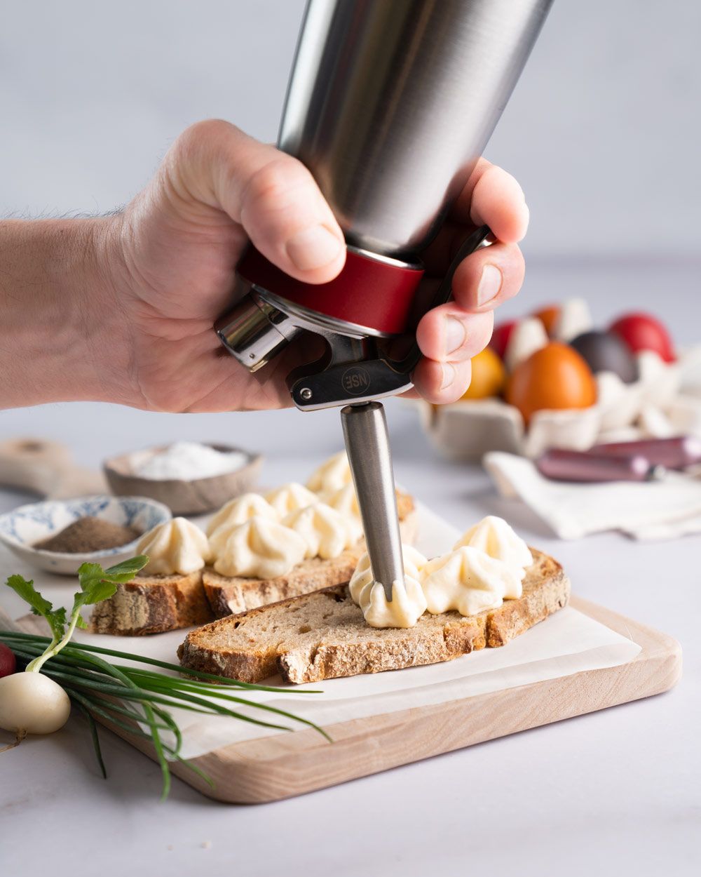 Jemand nutzt ein iSi Gourmet Whip um Eiaufstrich auf ein Brot zu sprühen, daneben liegt ein fertiges Brot und Kräuter. Im Hintergrund sind bunte Eier und zwei iSi Charge Kapseln zu sehen. 