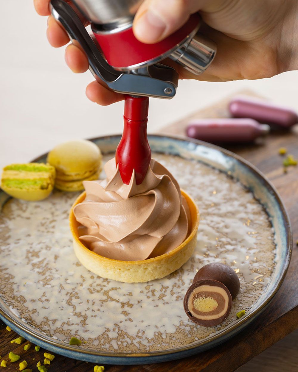 Jemand sprüht mit einem iSi Gourmet Whip Mozart Mousse auf ein Tarte, daneben  Mozartkugeln, iSi Charge Kapseln und Macarons.