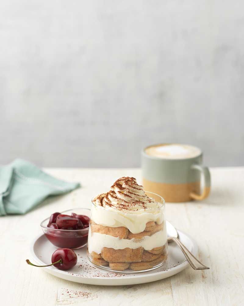 Ein Glas mit Tiramisu, daneben eine kleine Schlüssel mit Marmelade und eine Kirsche, im Hintergrund eine Tasse mit Kaffee.