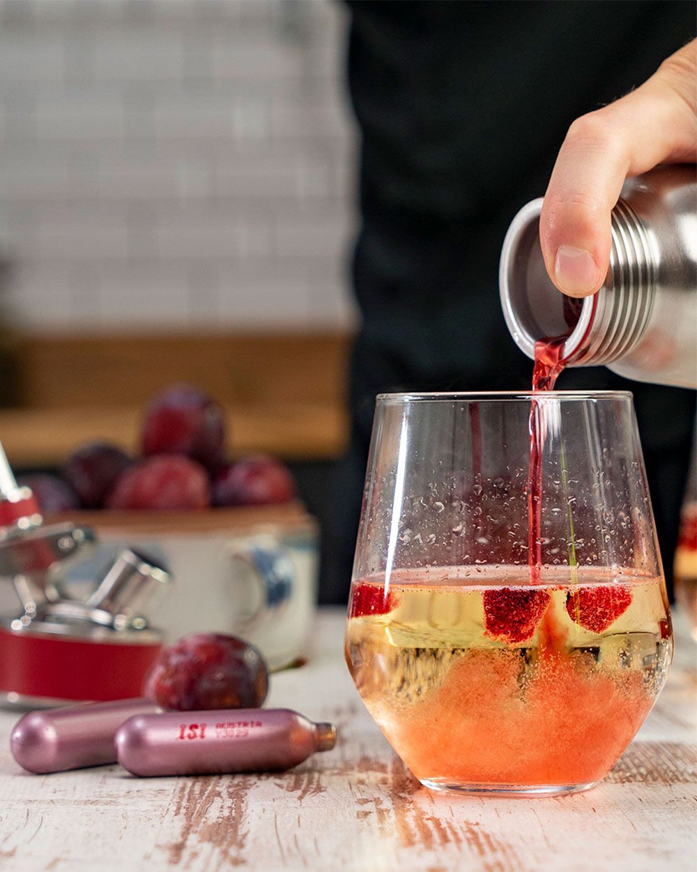 Jemand gießt Pflaumensirup aus einem iSi Gourmet Whip in ein Glas, daneben iSi Charge Kapseln. 