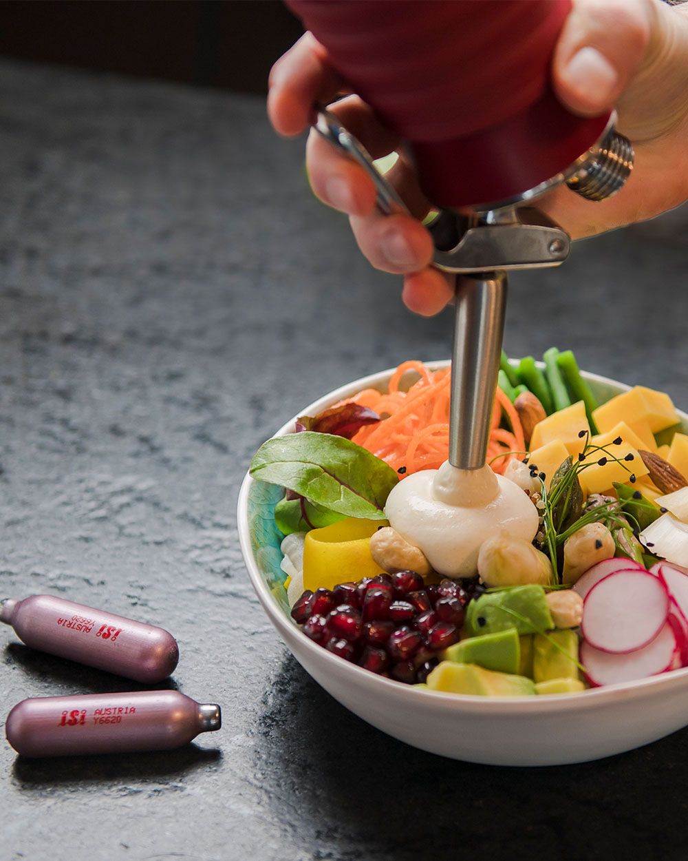 Jemand sprüht mit einem iSi Gourmet Whip vegane Sauce Hollandaise in eine Schüssel mit Gemüse, daneben liegen iSi Charge Kapseln.